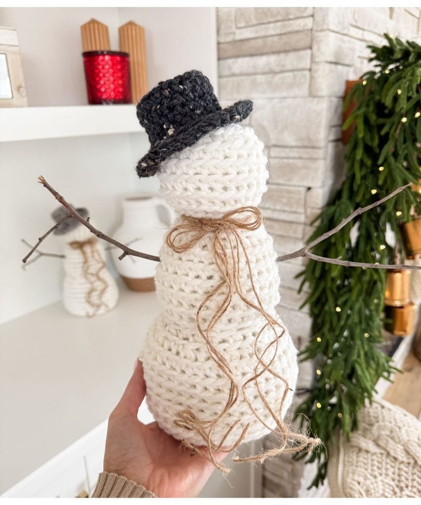 A hand holds a crocheted snowman with a black hat, brown string bow, and stick arms; festive decor and a crochet mushroom are visible in the background.
