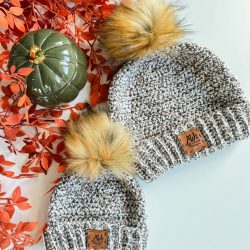Two gray knit beanies with faux fur pom-poms and leather tags are displayed next to a green ceramic pumpkin and orange leaves—perfect inspiration for your next project using the easiest crochet beanie or free crochet pattern.