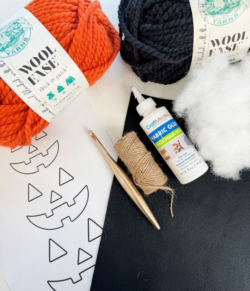 Orange and black yarn, a crochet hook, fabric glue, stuffing, twine, black felt, and pumpkin face templates are arranged on a white surface—everything you need to craft your own Crochet Jack-O-Lantern masterpiece.