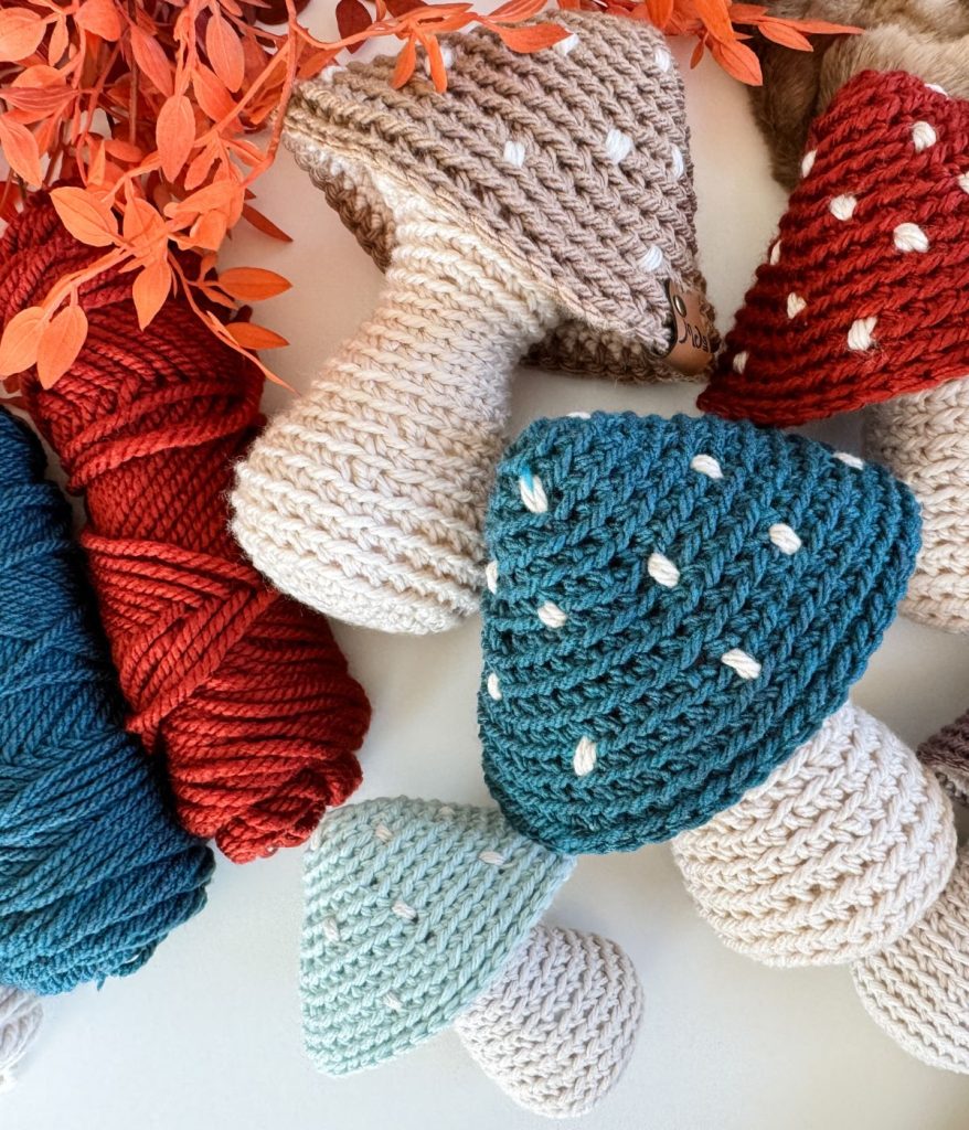 Crochet mushrooms in various colors are displayed with skeins of yarn and orange artificial leaves on a white surface.