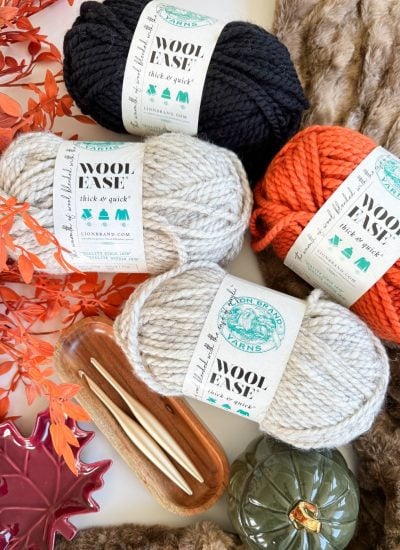Four skeins of Wool-Ease Thick & Quick yarn in black, white, gray, and orange are displayed with knitting needles, autumn leaves, a decorative pumpkin, and a faux fur fabric background—perfect for your next Crochet Jack-O-Lantern project.