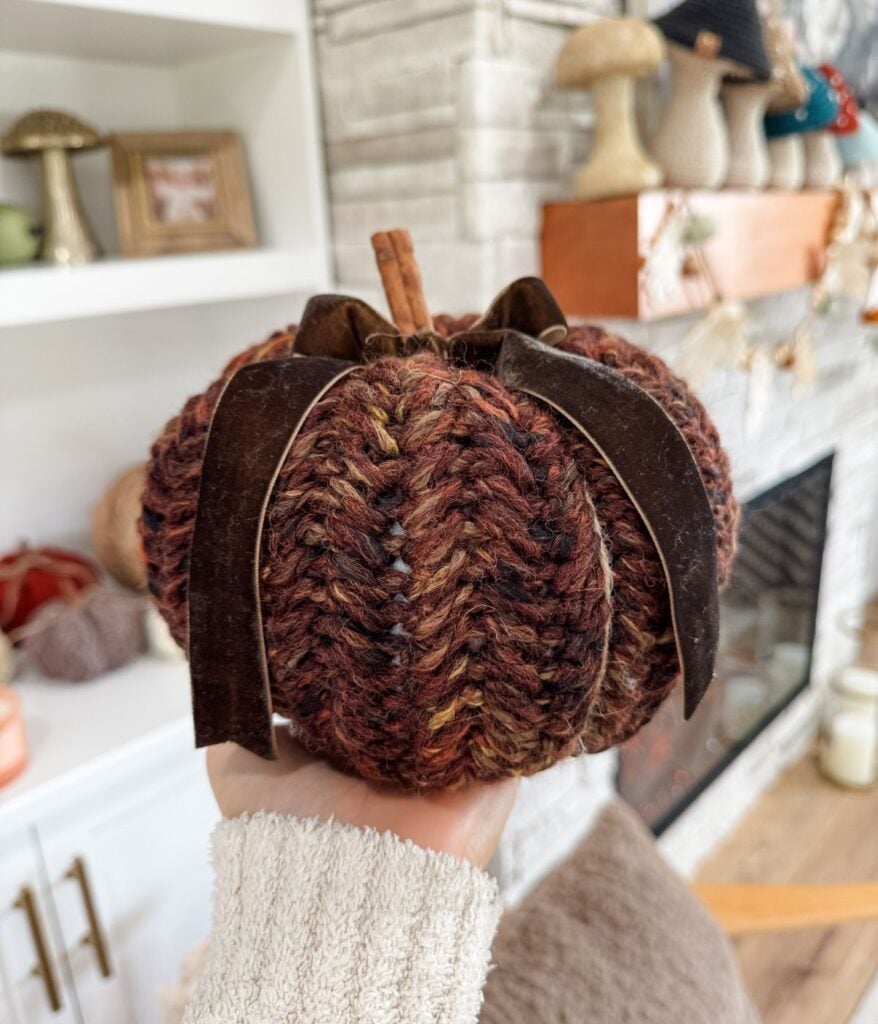 A hand holds a knitted Herringbone Pumpkin with a brown ribbon and cinnamon stick stem, in front of a cozy living room adorned with warm autumn decor.