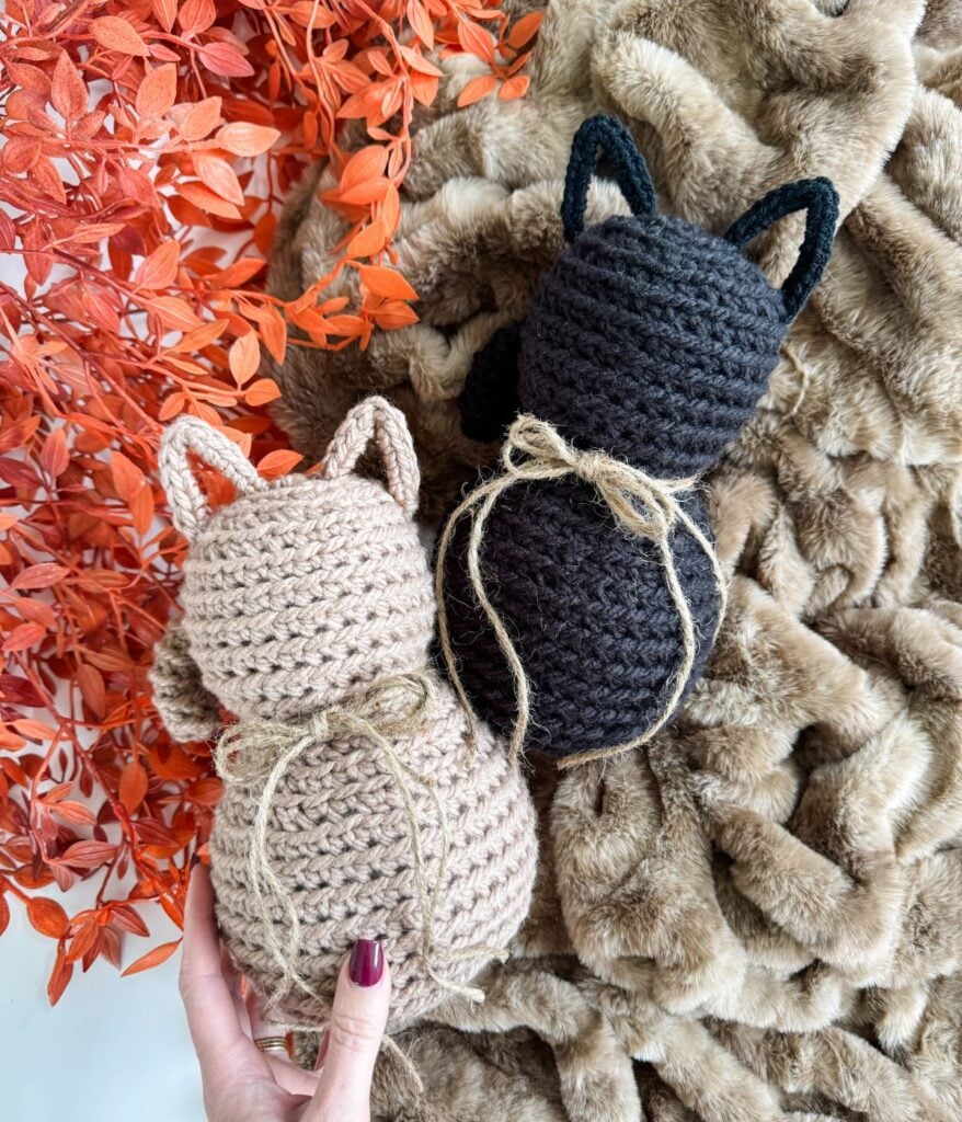 Two Crochet Cat figures, one beige and one black, each with twine tied around their necks, rest on a furry brown blanket beside orange foliage.