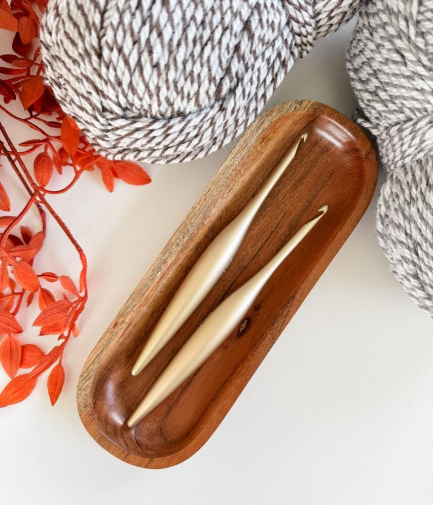 Two ergonomic crochet hooks on a wooden tray, surrounded by grey and white yarn balls and orange decorative leaves—the perfect setup for starting the easiest crochet beanie or following your favorite free crochet pattern.