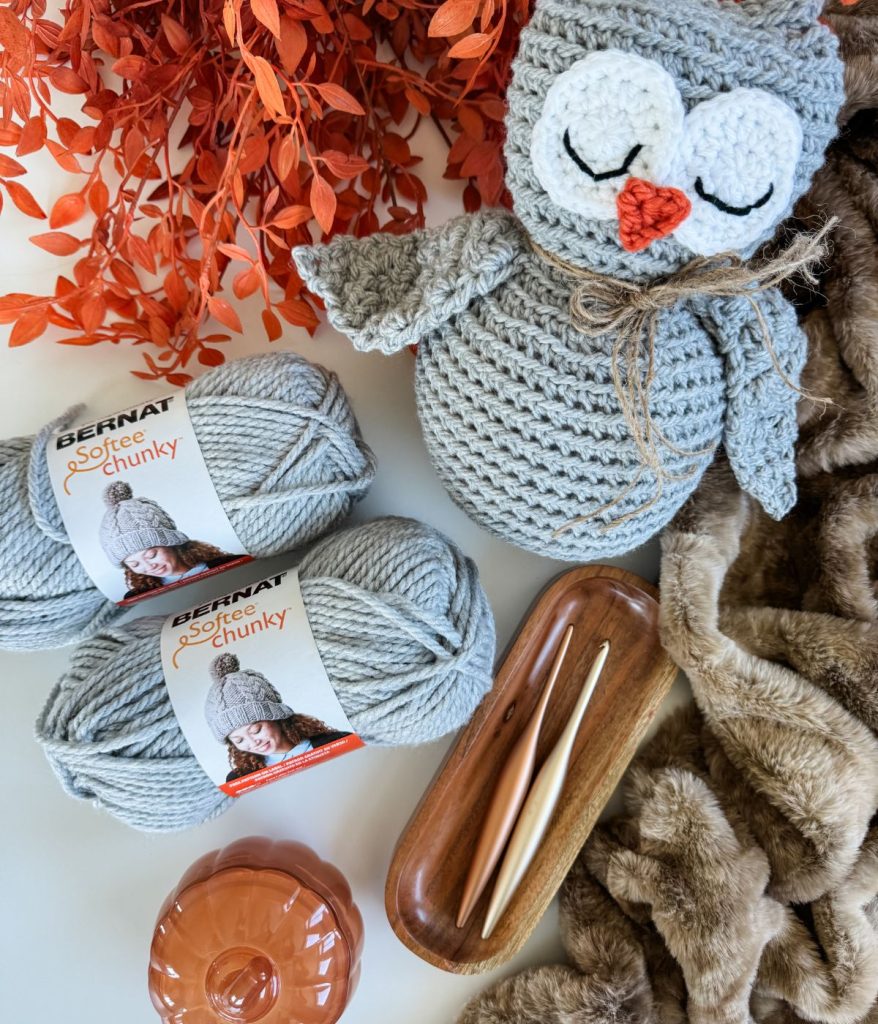 Gray crochet owl made with Bernat Softee Chunky yarn using the Owl Crochet pattern, displayed with two wooden crochet hooks on a wooden tray, faux orange foliage, and a brown faux fur blanket.