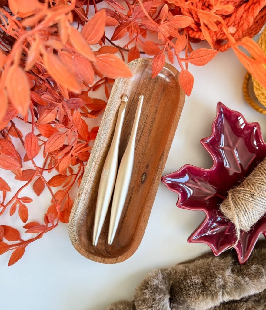 Two metallic crochet hooks rest in a wooden tray, surrounded by orange foliage, yarn, a Crochet Jack-O-Lantern, a burgundy leaf-shaped dish, twine, and a piece of faux fur.