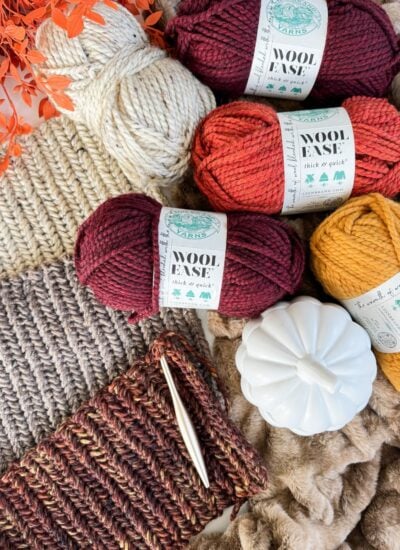Yarn skeins in red, yellow, and cream colors are placed on knitted fabric, alongside a crochet hook, faux pumpkin, herringbone pumpkin, and orange foliage.