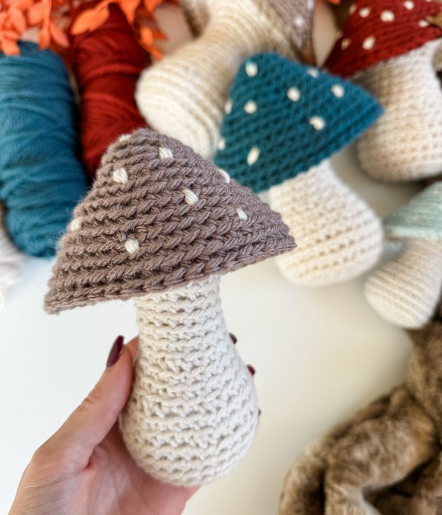 A hand holds a Crochet Mushroom with a beige stem and a brown cap with white spots. Other crocheted mushrooms and yarn balls are visible in the background.