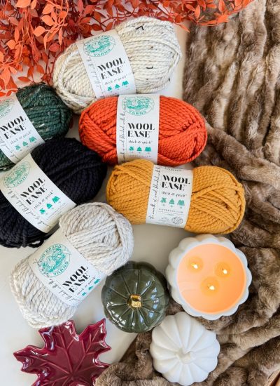 Five skeins of yarn in green, cream, orange, black, and yellow are arranged with a faux fur blanket, decorative pumpkins, candles, orange leaves, a ceramic leaf dish, and a charming Crochet Jack-O-Lantern.