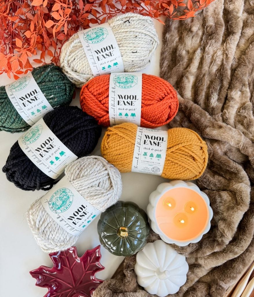 Five skeins of yarn in green, cream, orange, black, and yellow are arranged with a faux fur blanket, decorative pumpkins, candles, orange leaves, a ceramic leaf dish, and a charming Crochet Jack-O-Lantern.