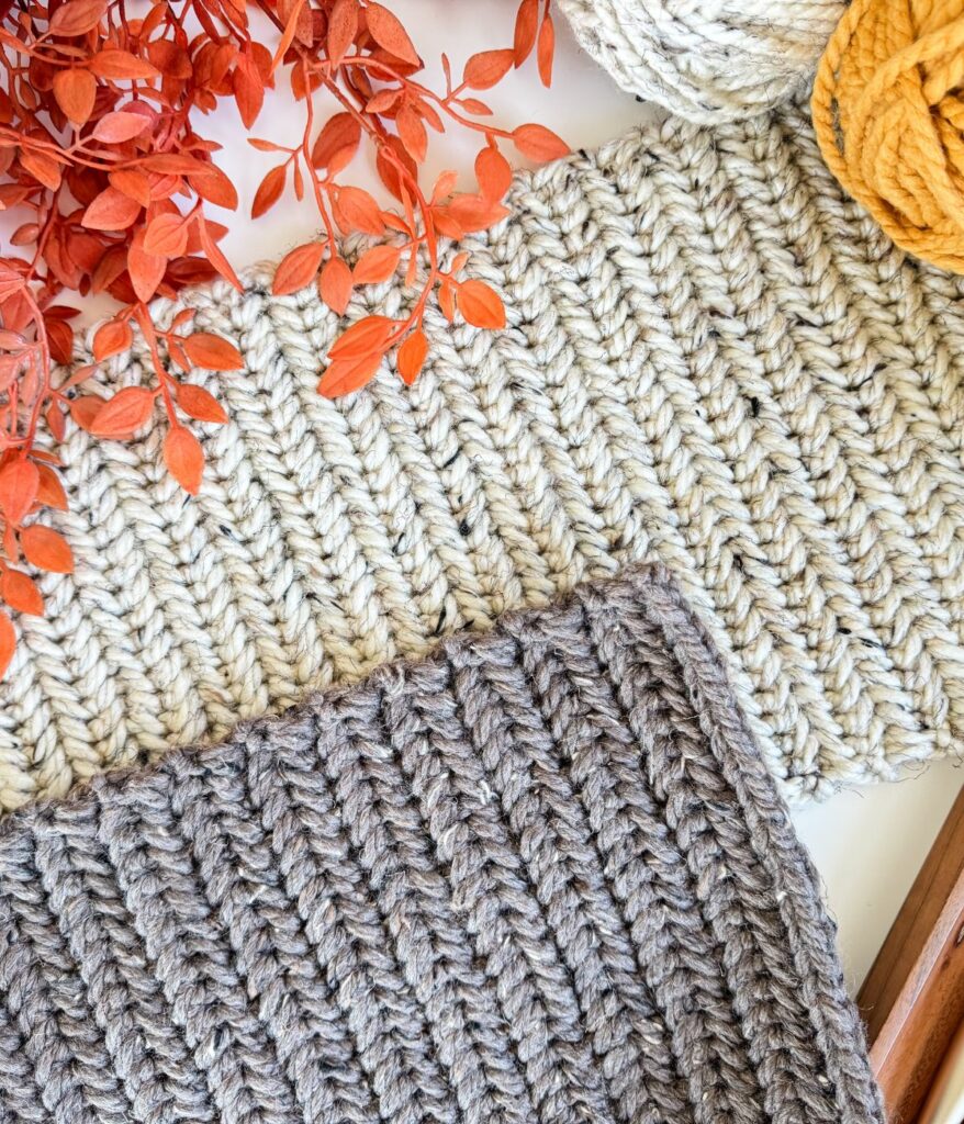 Close-up of two knitted fabric pieces, one beige and one gray, placed next to orange artificial leaves, mustard yellow yarn, and a cozy Herringbone Pumpkin.