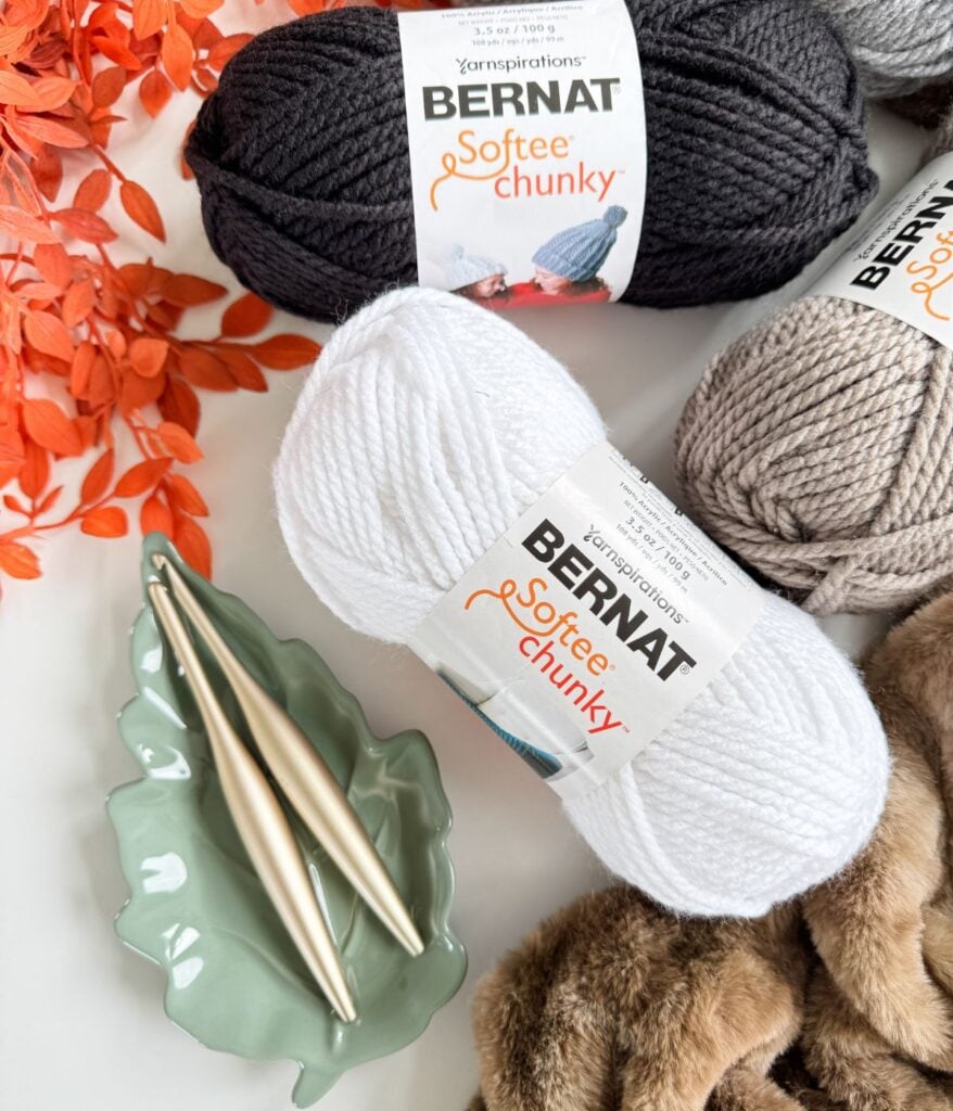 Three skeins of Bernat Softee Chunky yarn in black, white, and beige—perfect for crafting a Crochet Cat—rest with two knitting needles on a green leaf-shaped dish, surrounded by decorative orange foliage and faux fur trim.