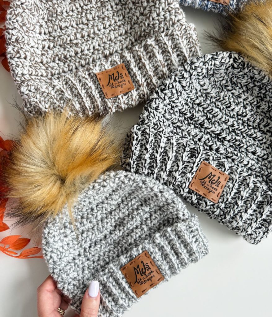 Three knitted beanies with leather tags, including styles inspired by the easiest crochet beanie patterns, two with faux fur pom-poms, displayed on a white surface with an orange ribbon partly visible.