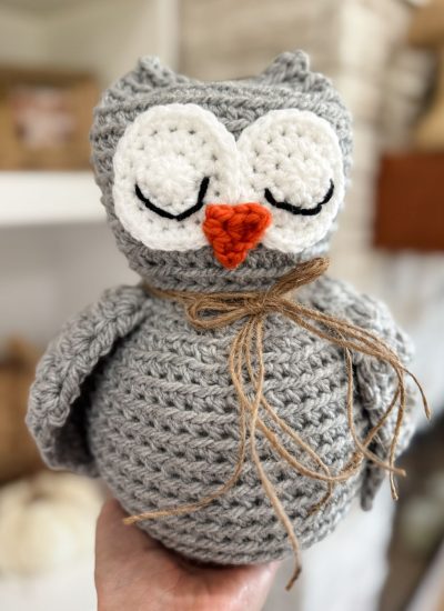 A hand holds a crocheted gray owl with closed eyes, an orange beak, and a twine bow around its neck—crafted using an adorable Owl Crochet pattern.
