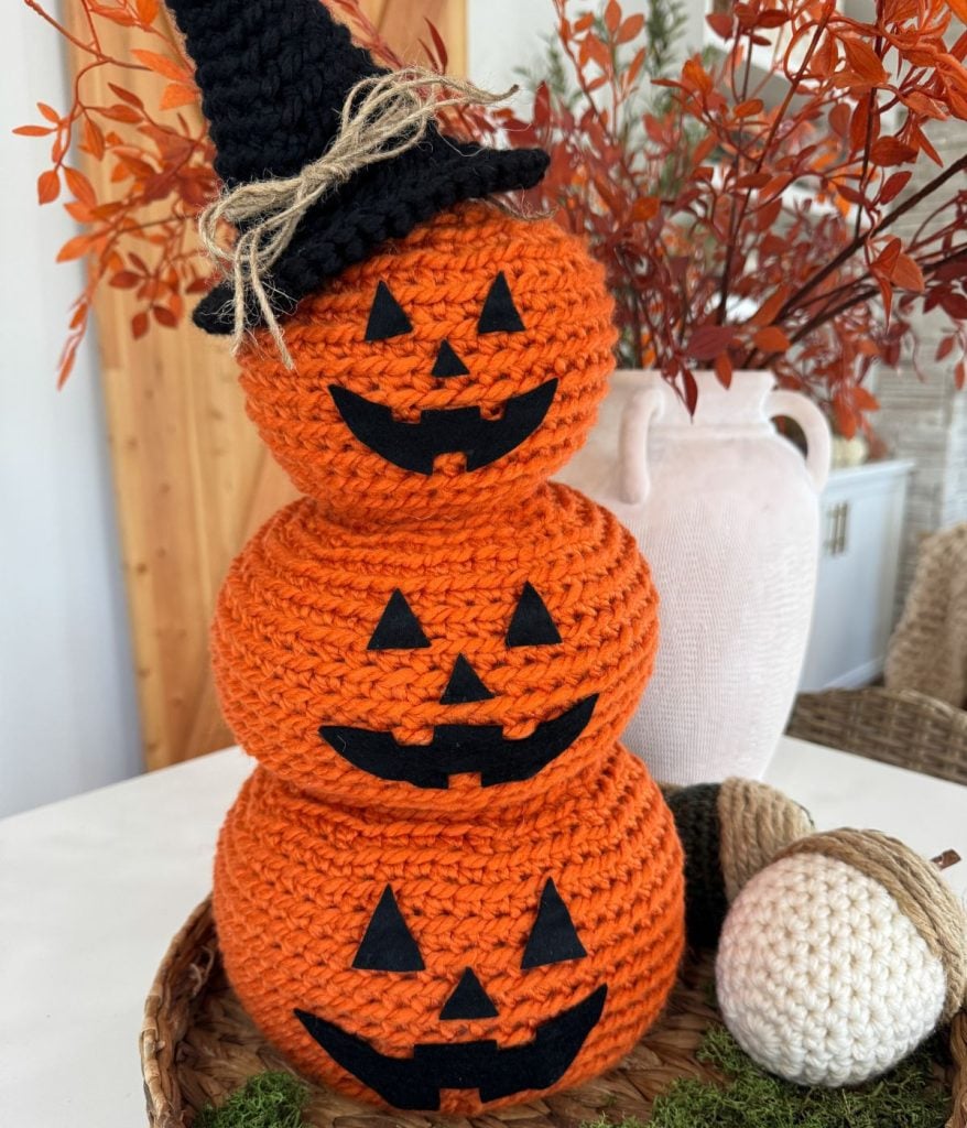 A stack of three Crochet Jack-O-Lantern pumpkins with black jack-o'-lantern faces, topped with a black witch hat, displayed on a tray with festive fall decor.