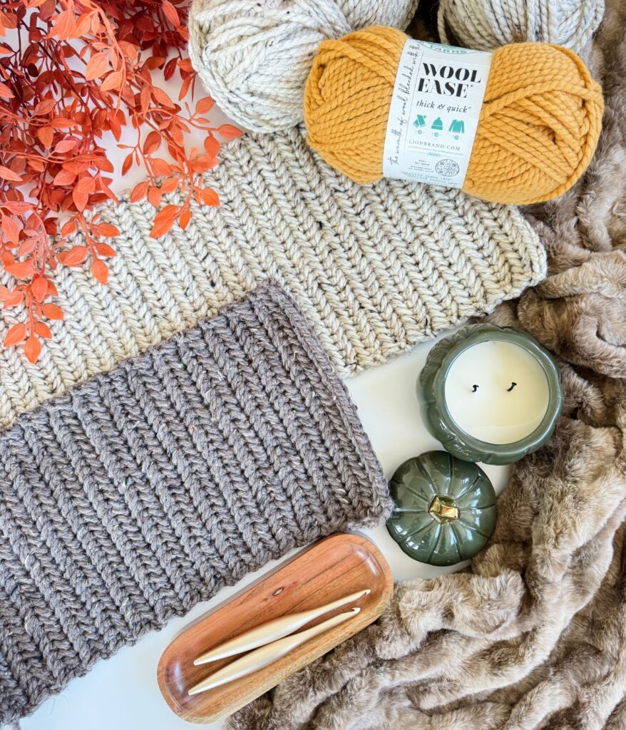 Two knitted swatches, skeins of yarn, a candle, two crochet hooks in a wooden tray, faux fur fabric, and orange decorative leaves are arranged flat on a surface alongside a charming Herringbone Pumpkin.