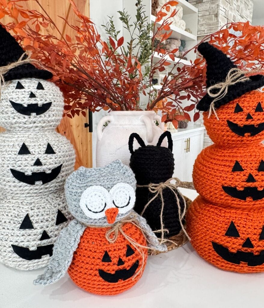 Crocheted Halloween decorations featuring a crochet cat, stacked pumpkins, a witch hat, an owl, and autumn foliage arranged in a white vase.