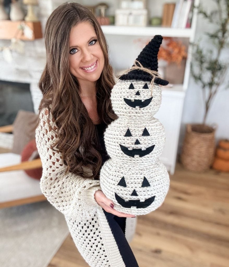 Woman holding a Crochet Jack-O-Lantern decoration featuring three stacked jack-o'-lanterns with a black witch hat, standing in a cozy, decorated living room.