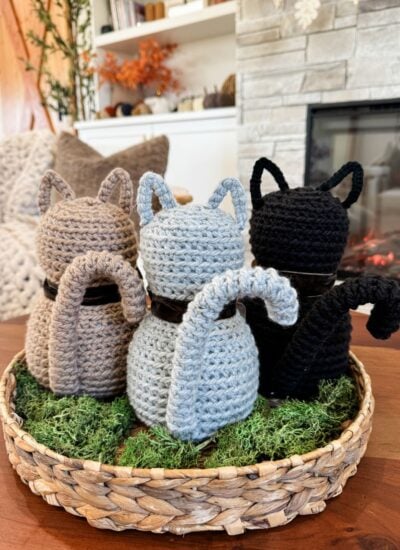 Three Crochet Cat figures in brown, gray, and black sit on a bed of green moss in a woven basket atop a wooden table near a cozy fireplace.