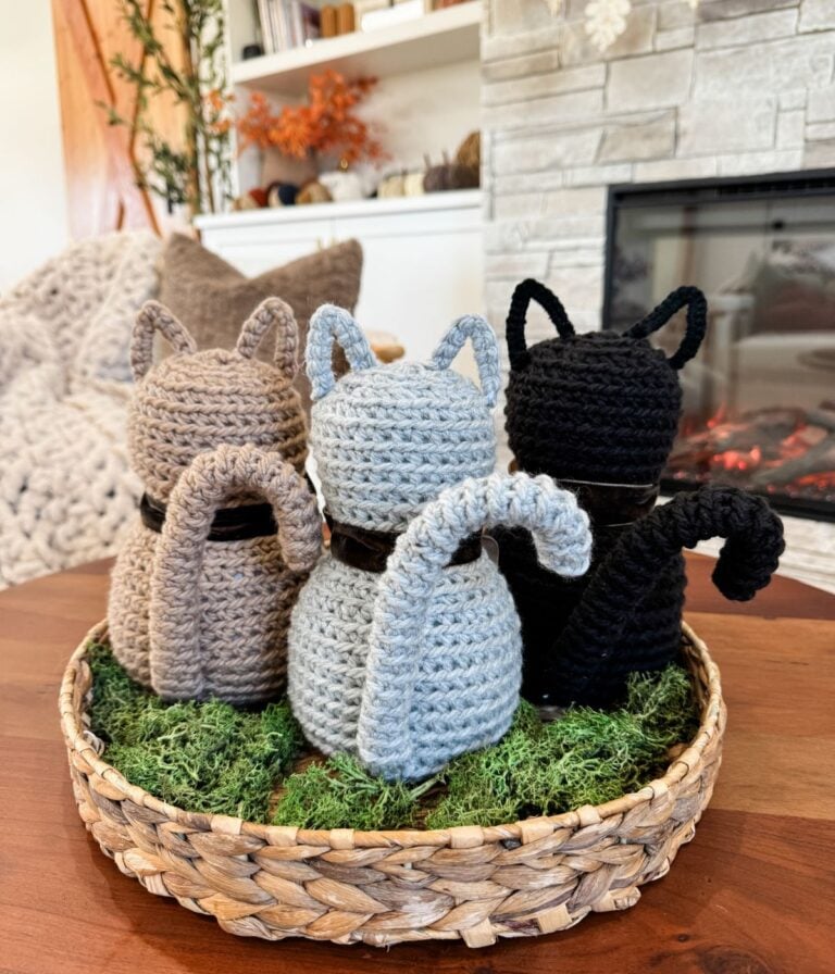 Three Crochet Cat figures in brown, gray, and black sit on a bed of green moss in a woven basket atop a wooden table near a cozy fireplace.