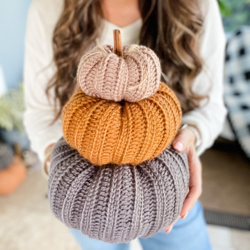A person holds three stacked, knitted pumpkins in gray, orange, and beige beside a charming crochet mushroom.