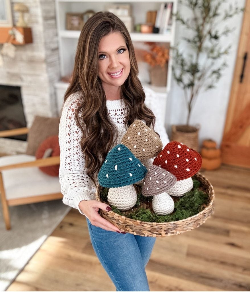 A woman in a white sweater holds a basket filled with colorful crochet mushrooms, standing in a cozy, well-lit living room.