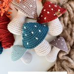 A group of Crochet Mushrooms in red, blue, and teal with beige stems displayed on a surface with yarn and leaves nearby. Text at the bottom reads "Rustic Farmhouse Mushrooms.