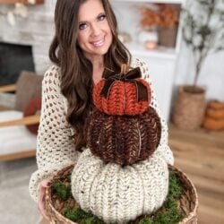 A woman in a cream knit sweater holds a basket with three stacked, knitted Herringbone Pumpkins in autumn colors, placed on a bed of moss.