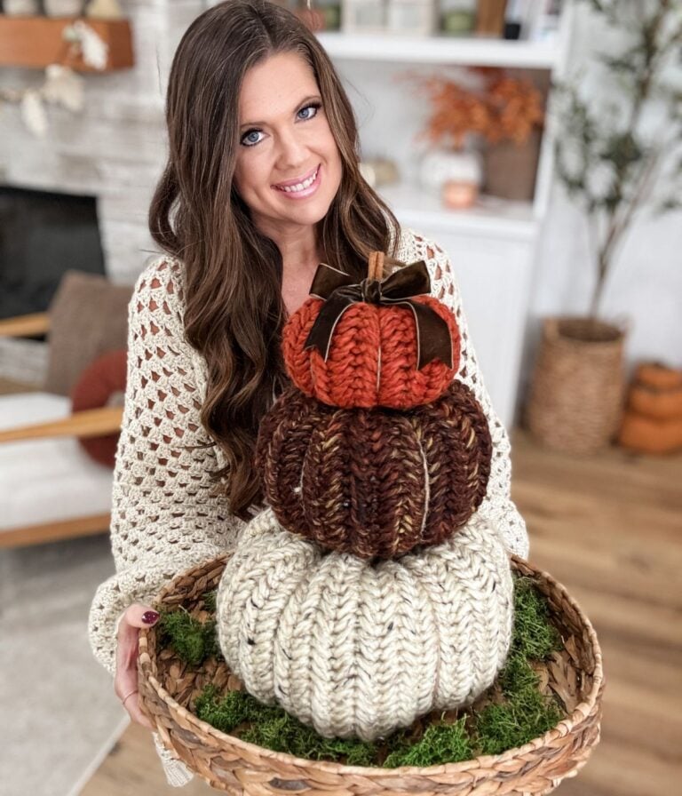 A woman in a cream knit sweater holds a basket with three stacked, knitted Herringbone Pumpkins in autumn colors, placed on a bed of moss.
