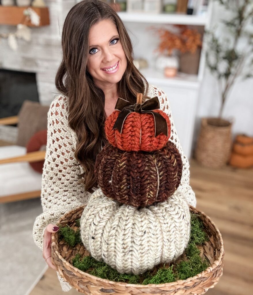 A woman in a cream knit sweater holds a basket with three stacked, knitted Herringbone Pumpkins in autumn colors, placed on a bed of moss.