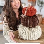 A woman holds a wooden tray with three crocheted pumpkins, including a charming Herringbone Pumpkin, stacked in cream, brown, and orange hues.