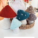 Five Crochet Mushroom figures with textured stems and colorful caps are displayed on a wicker tray, with a white vase of red foliage in the background.