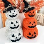 Two Crochet Jack-O-Lantern stacks, one white and one orange, each with smiling faces and black hats, are displayed on a table adorned with autumn decor in the background.