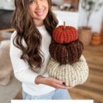 A woman with long brown hair holds a stack of three Herringbone Pumpkins in orange, brown, and cream. Text below reads: "Chunky Herringbone Pumpkins. Free Crochet Pattern With Video Tutorial.