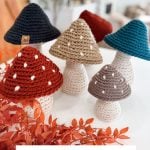 A group of Crochet Mushroom decorations in various colors and sizes is displayed on a white surface, with orange foliage in the foreground.