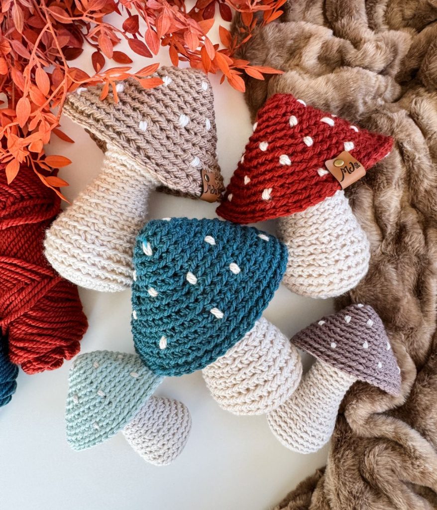 Five Crochet Mushrooms with different colored caps—green, blue, red, purple, and beige—are arranged on a furry surface with red yarn and faux leaves beside them.