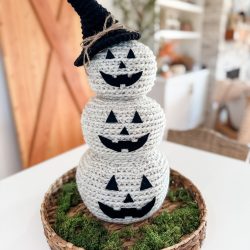 A Crochet Jack-O-Lantern snowman with three stacked pumpkin faces and a black witch hat sits on a woven tray with green moss.