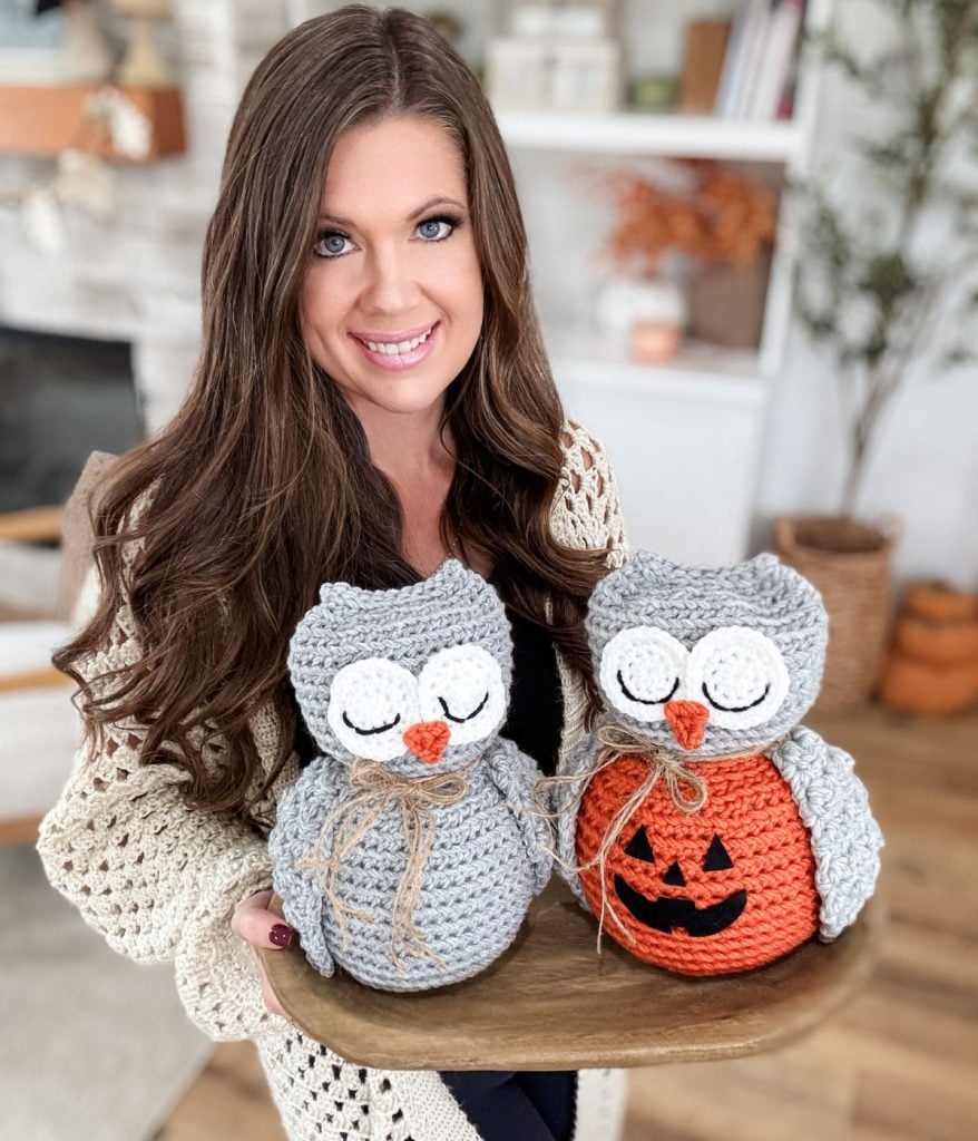 A woman holds a wooden tray with two adorable creations made from an Owl Crochet pattern—one gray and one orange with a jack-o'-lantern face—in a cozy living room.