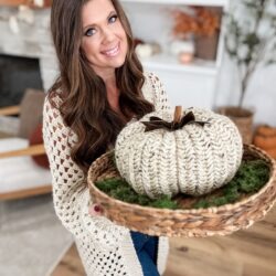 Woman holding a woven basket with a Herringbone Pumpkin and green moss, standing in a cozy, decorated living room.
