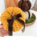 A hand holds a chunky Herringbone Pumpkin crocheted in yellow, featuring a velvet ribbon and cinnamon stick stem. Two more Herringbone Pumpkins rest in a basket behind. Text below reads "Chunky Herringbone Pumpkins.