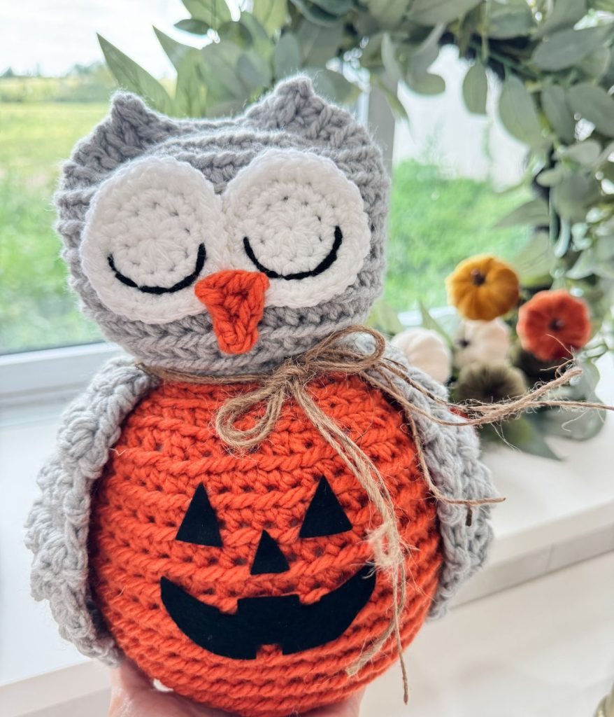 A crocheted owl with a gray head and wings sits on an orange pumpkin body with a black Jack-o'-lantern face, crafted from an adorable Owl Crochet pattern, in front of a window with greenery and decorations.