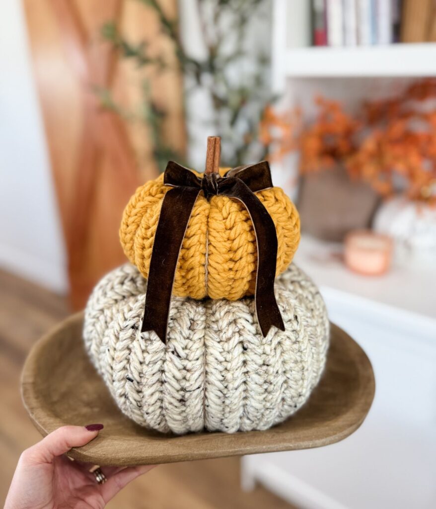 A hand holds a wooden tray with two stacked knitted pumpkins—a yellow Herringbone Pumpkin with a ribbon on top and a cream-colored one beneath.