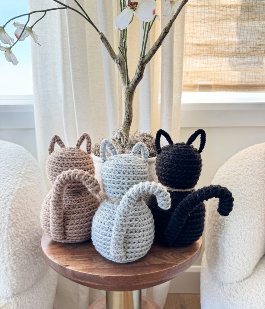 Three Crochet Cat figures in beige, gray, and black sit side by side on a round wooden table in front of a potted plant.