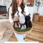 A woman holding a woven tray displays three adorable Crochet Cat figures in brown, gray, and black, standing in a cozy, rustic living room.