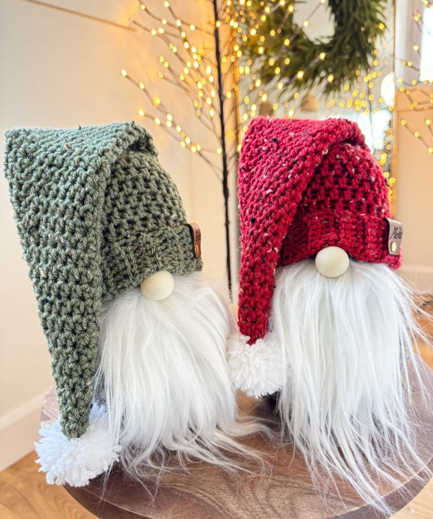 Two gnome figurines with white beards and large hats, one green and one red, sit on a wooden surface beside a crochet mushroom, with festive lights glowing softly in the background.