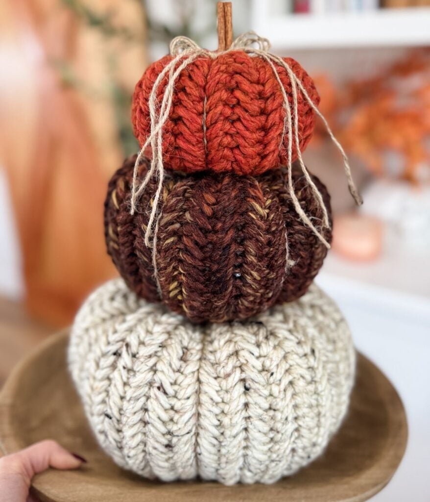 Three herringbone pumpkins in orange, brown, and cream are stacked on a wooden plate, each topped with twine bows, set against a blurred indoor background.