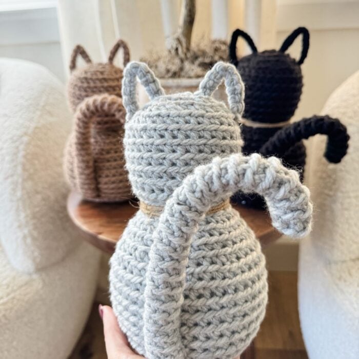 A hand holds a Crochet Cat in gray with a curled tail; two more crocheted cats in brown and black sit on a table in the background.