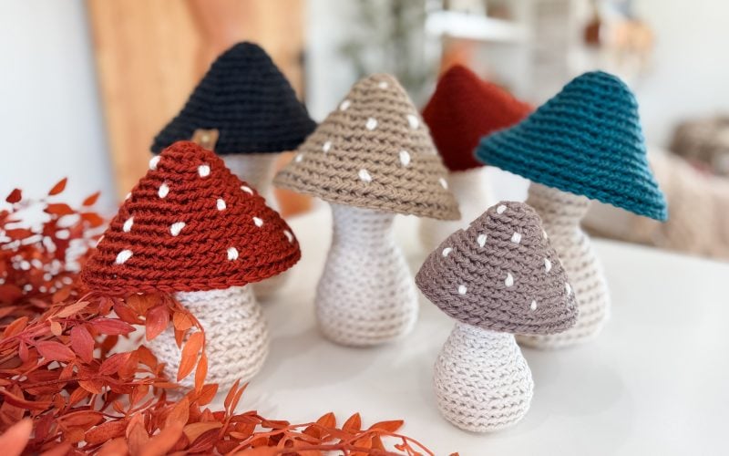 Six crocheted mushrooms in various colors and sizes are displayed on a white surface, with orange foliage in the foreground and a blurred indoor background.