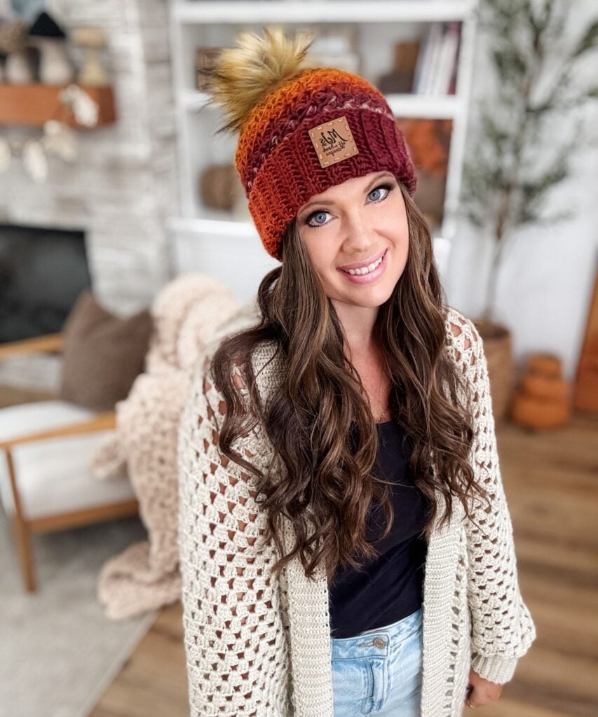 Woman with long brown hair wearing a Chunky Suzette Stitch Beanie with a pom-pom and a cream cardigan, standing indoors in a cozy, decorated living room.