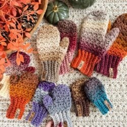 Several pairs of crocheted fingerless gloves and mittens in various colors are arranged on a textured surface, accented by autumn-themed decorations and a lit candle nearby.